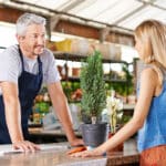 Employé d'une jardinerie portant un tablier bleu discutant avec une cliente blonde près d'un comptoir, avec des plantes et des fournitures en arrière-plan.